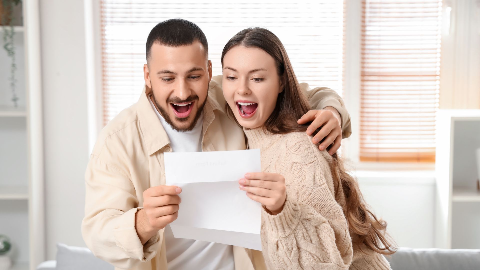 Smiling couple reading document with excitement indoors, sunlight through blinds, suggesting rental approval or lease success.