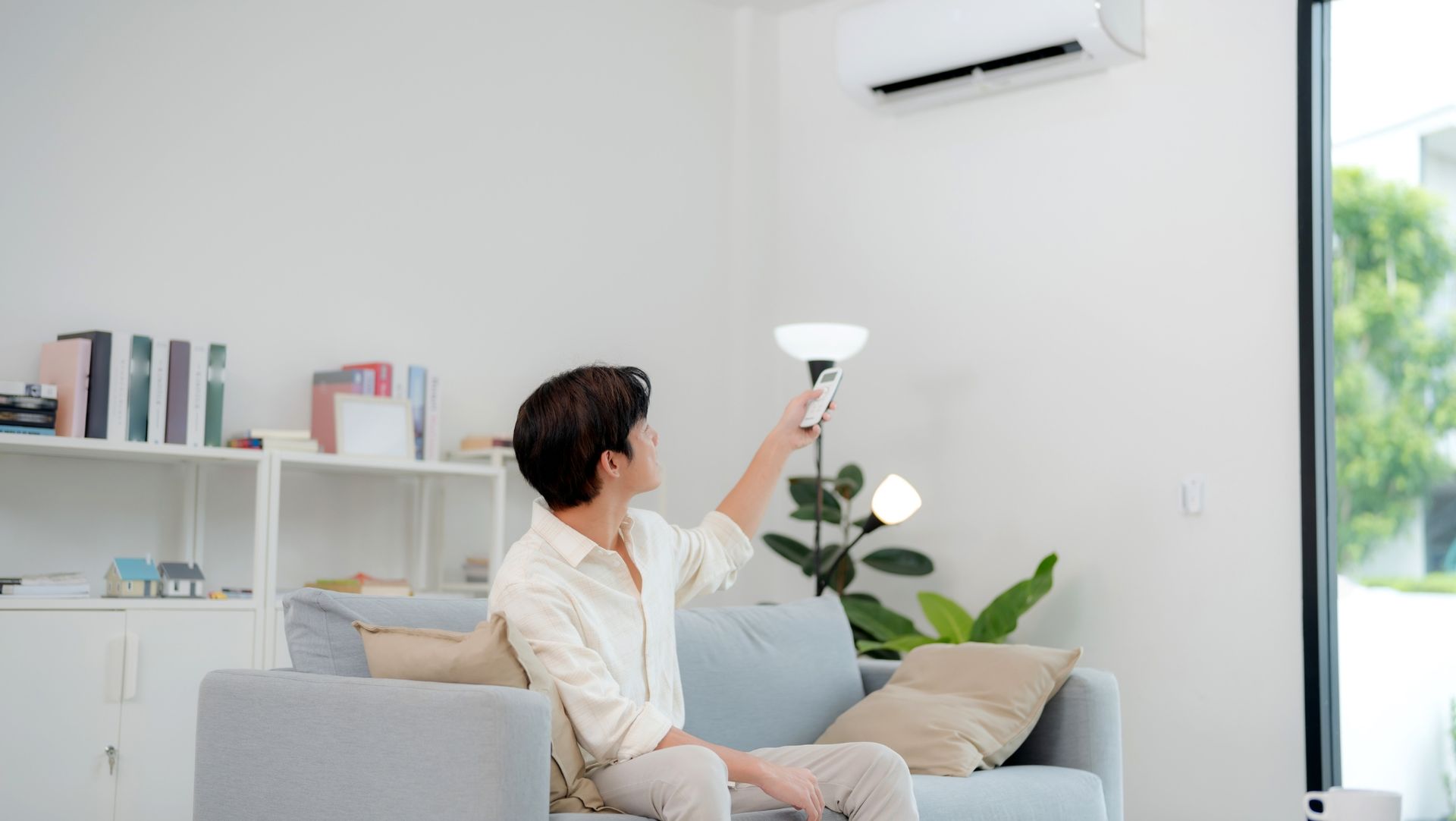 Person on gray sofa using remote to adjust wall-mounted AC in modern living room with plants, bookshelves, and floor lamp.
