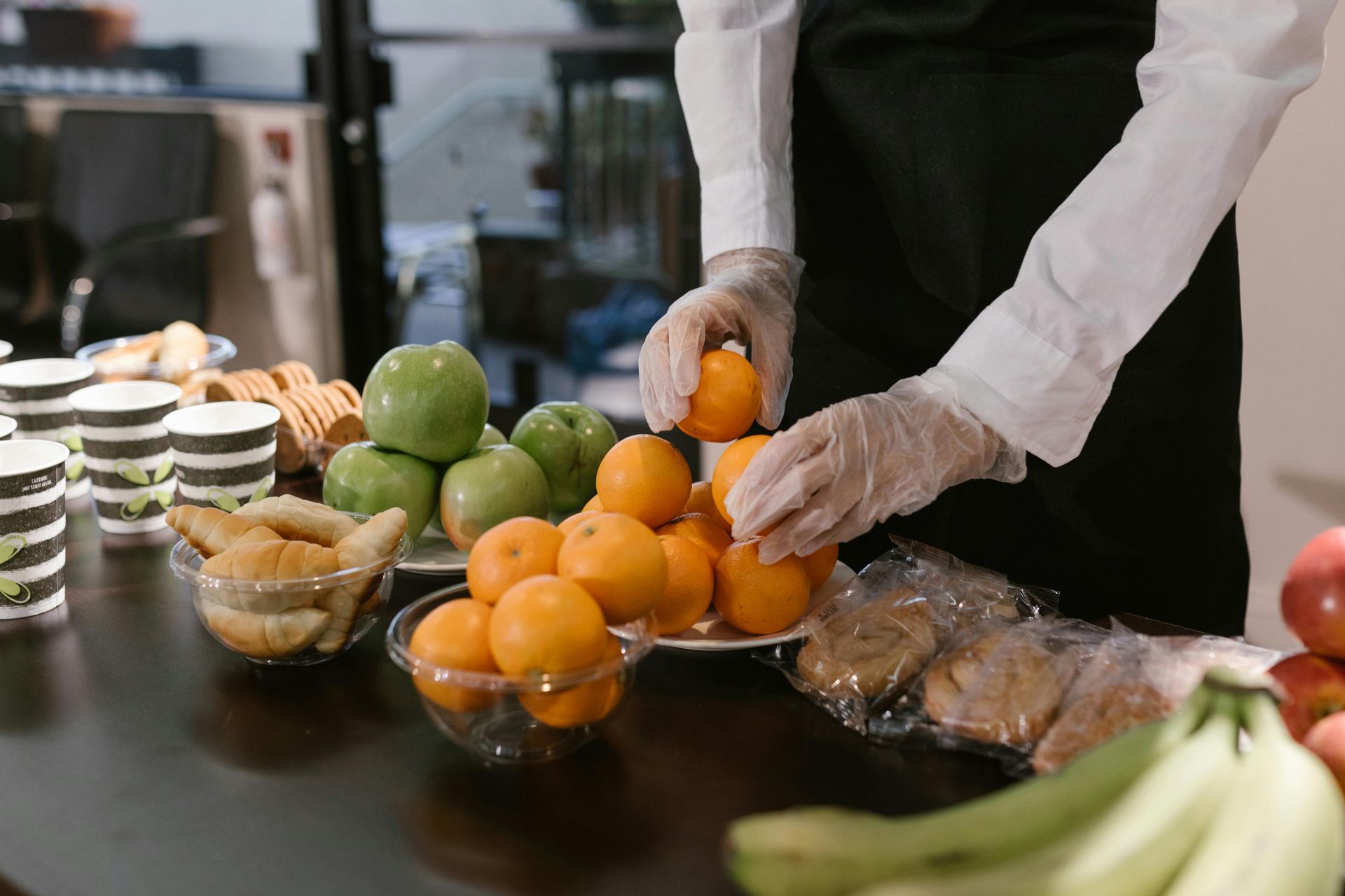 Gloved hands arranging oranges and apples on a buffet table with pastries and bananas nearby