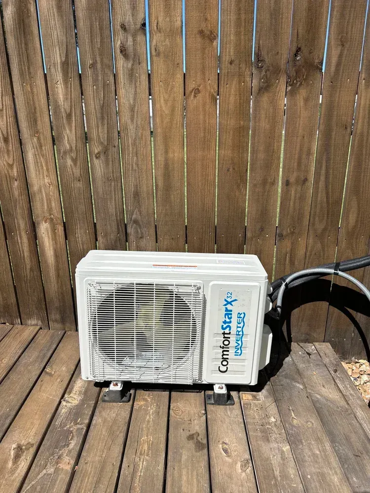 Outdoor air conditioner unit on a wooden deck in front of a weathered fence.