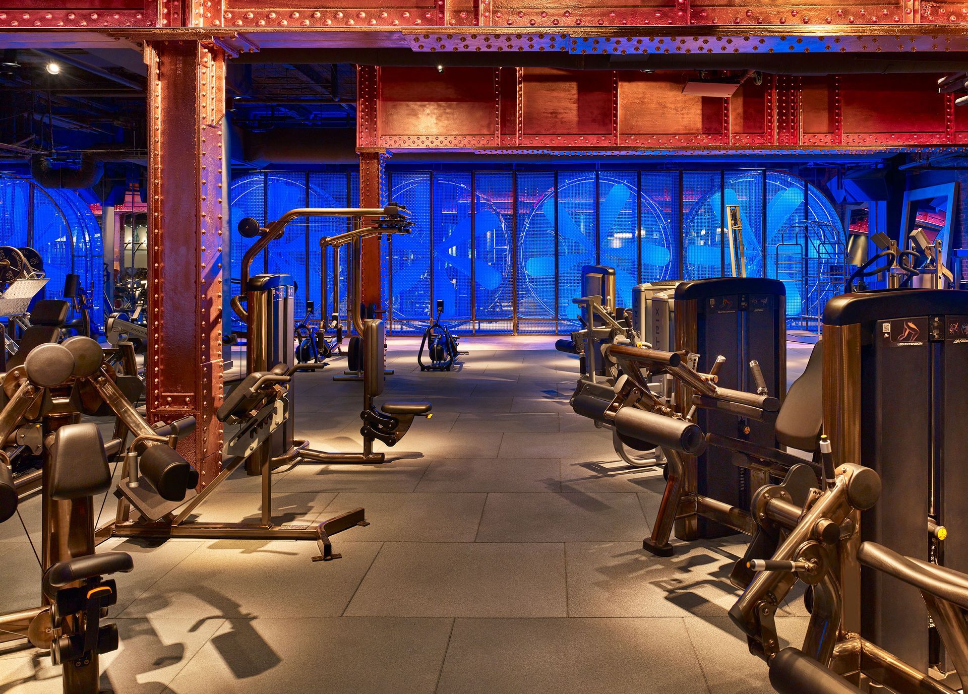 Gym interior with weight machines, brown steel structure, blue lighting.