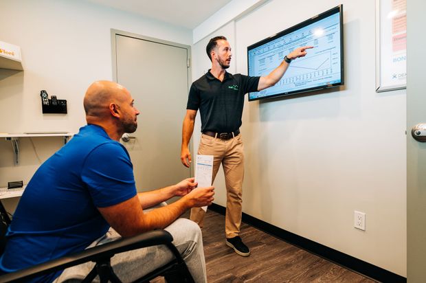 Man points at a screen, explaining to another man seated in a room with a whiteboard, discussing a diagram.