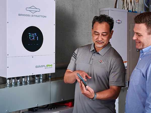 Two men examining a SimpliPhi battery system. One points at a phone, the other looks on in a basement.