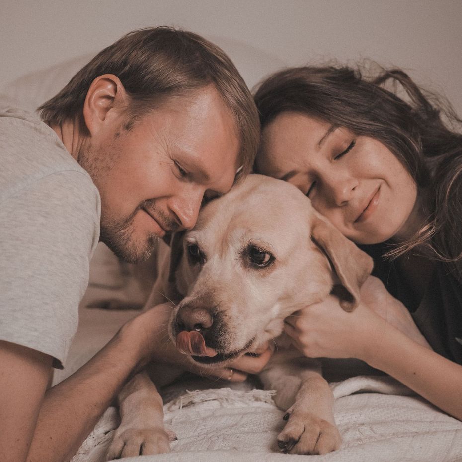 Couple cuddling a dog on a bed, all three resting close with gentle smiles