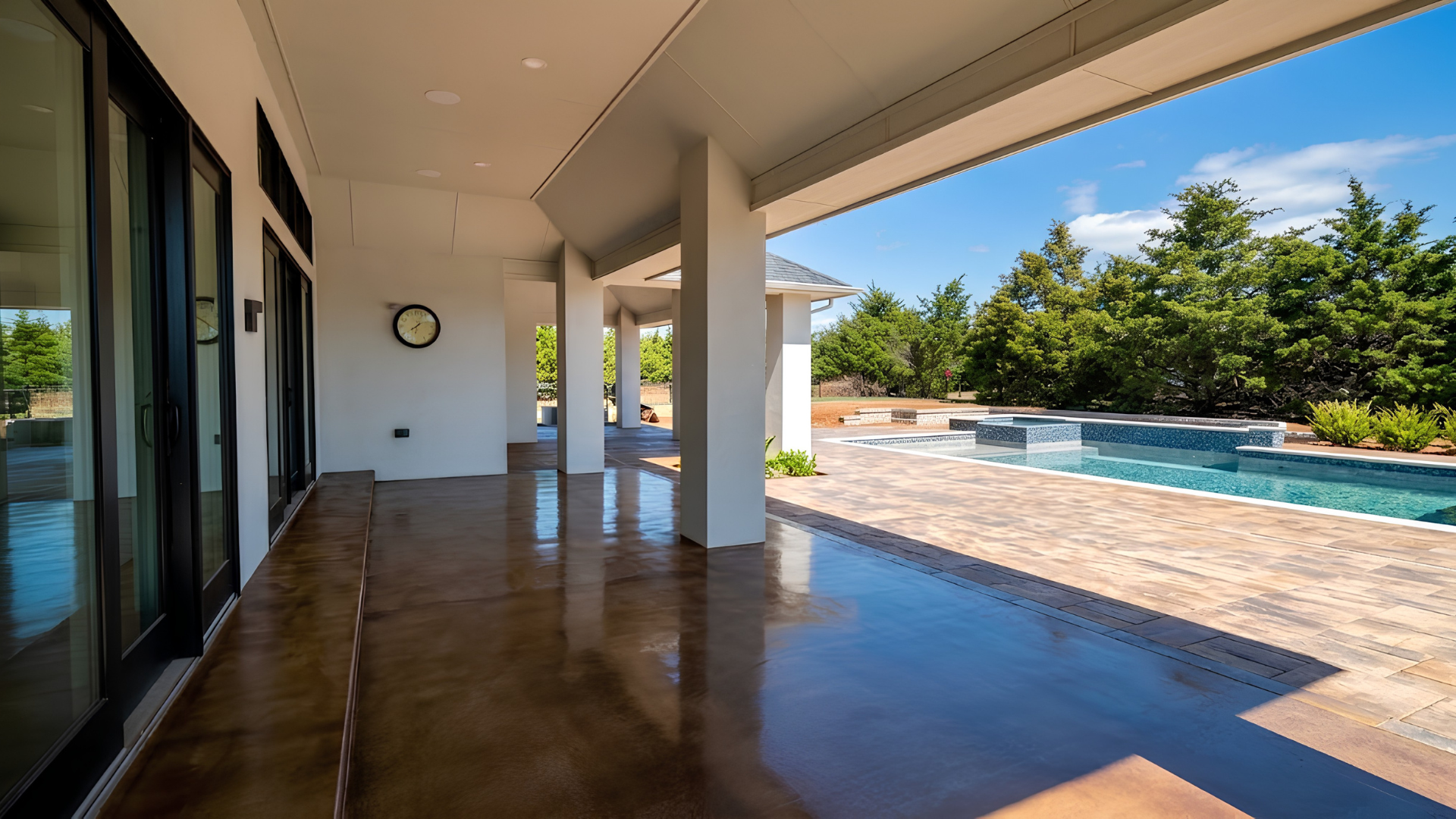 Covered patio beside a sunny backyard pool, with glass doors and blue water surrounded by trees.