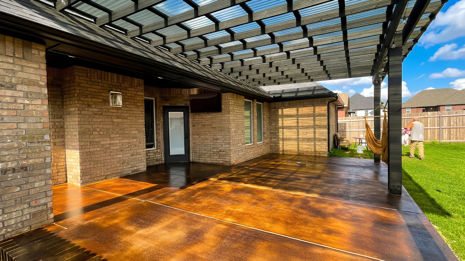 Covered brick patio with dark wood deck, glass pergola roof, and a view of green lawn and blue sky.