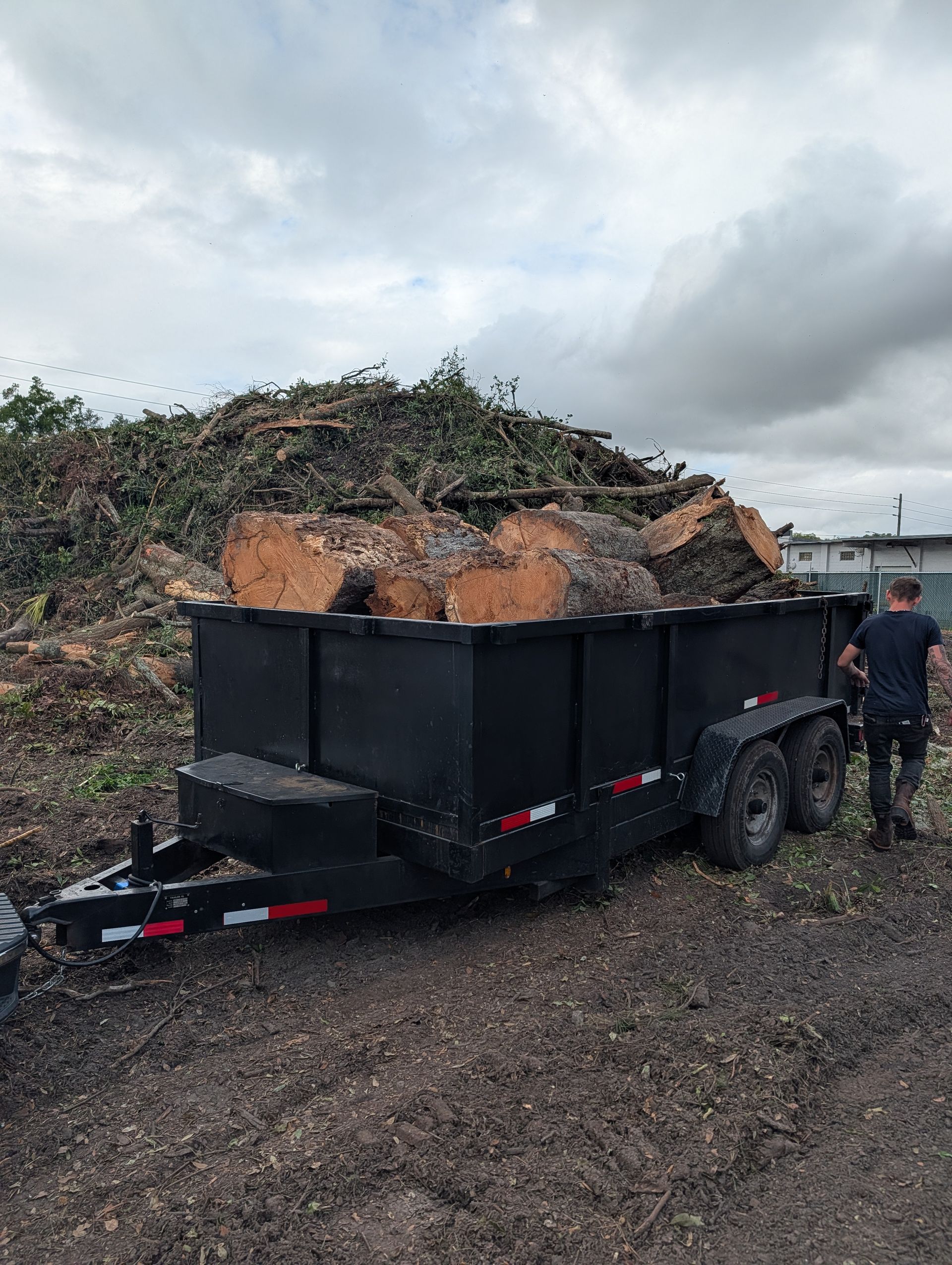 Black dump trailer loaded with soil and brush, parked on gravel under cloudy skies, with a person beside it
