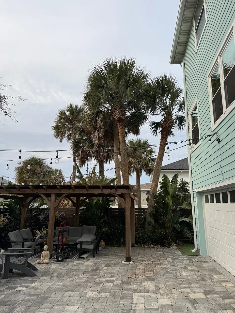 Stone patio with pergola and outdoor seating beside a pale blue house, with tall palm trees in the yard.
