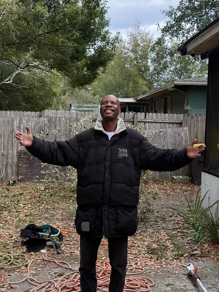 Person standing outdoors with arms raised, in a black coat near a fenced yard and house.