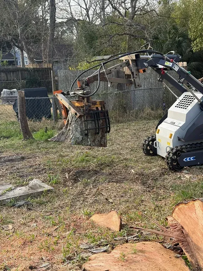 Small excavator with grapple lifting a burning stump in a brushy yard.