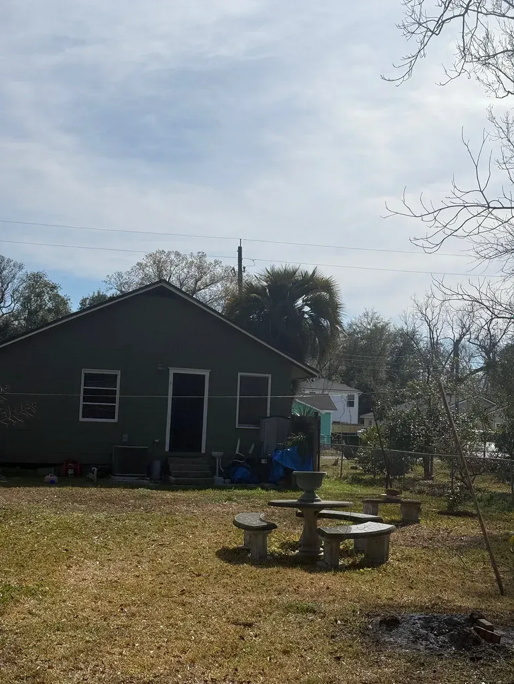Small dark house in a yard with a picnic table, fire pit, and trees under a cloudy sky.