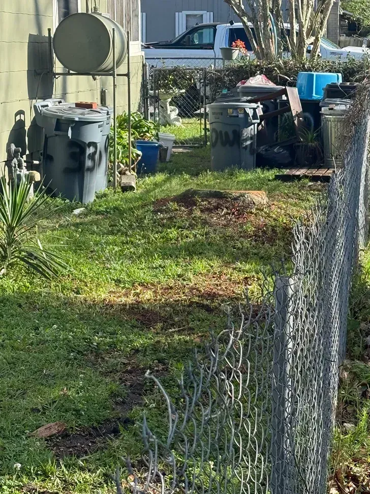 Chain-link fence beside a grassy yard with utility equipment and bins near a house.