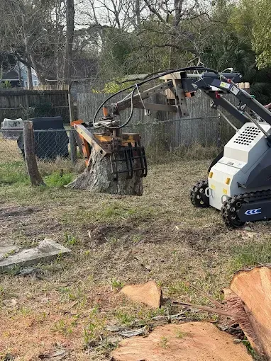 A compact excavator arm cuts a tree stump in a grassy yard with fallen logs nearby.