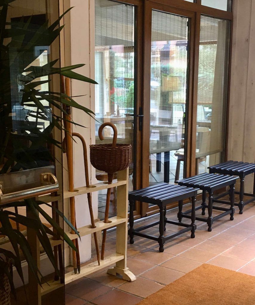 Interior with a plant, walking sticks, woven basket, and three small stools near a glass door.
