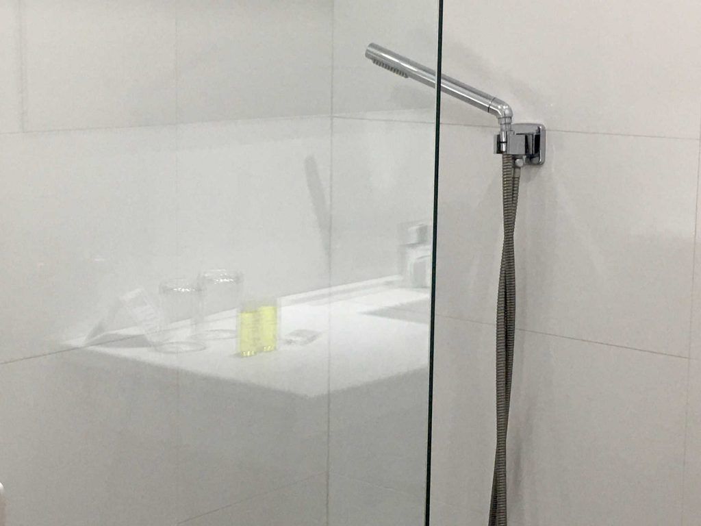 Shower with glass door, white tiled walls. Chrome showerhead and arm. Sink and toiletries visible through the glass.