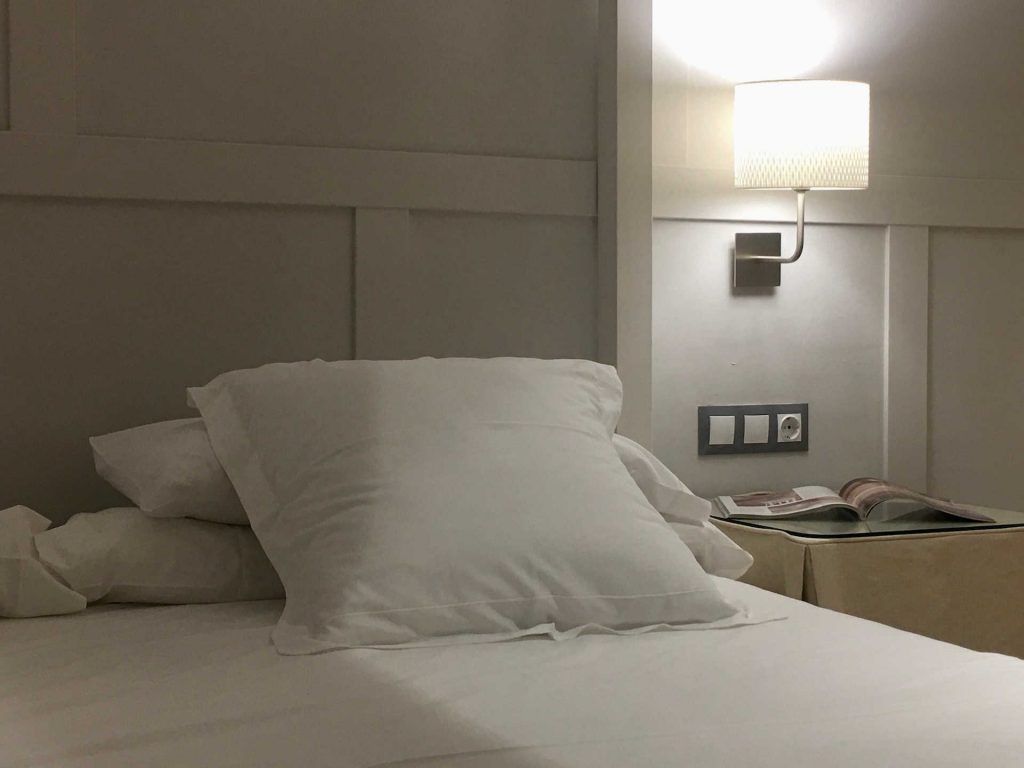 Bed with white pillows and sheets, a bedside lamp, and a nightstand with magazines in a hotel room.