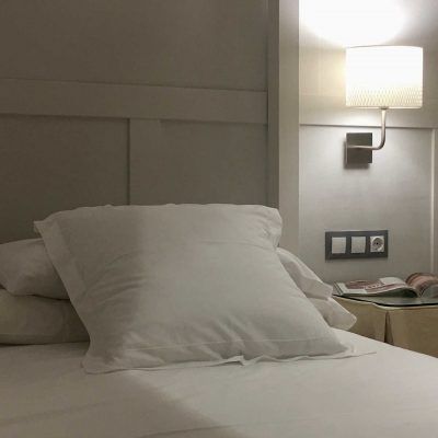 Close-up of a bed with white pillows and a lit wall lamp. A nightstand and power outlets are visible.