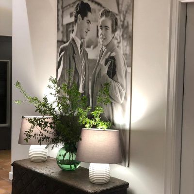 Black and white photograph of two men on a wall. Green vase with greenery and two lamps on a wooden chest in front.