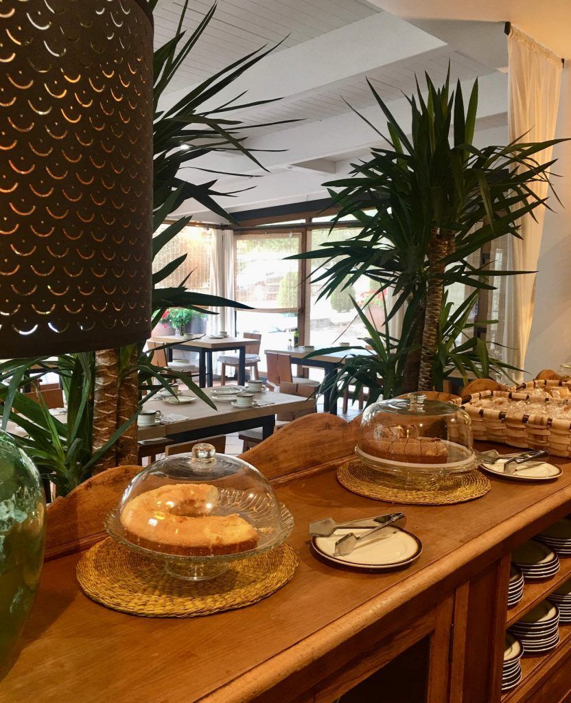 Wooden counter with cakes under glass domes, in front of a restaurant with tables and palm trees.