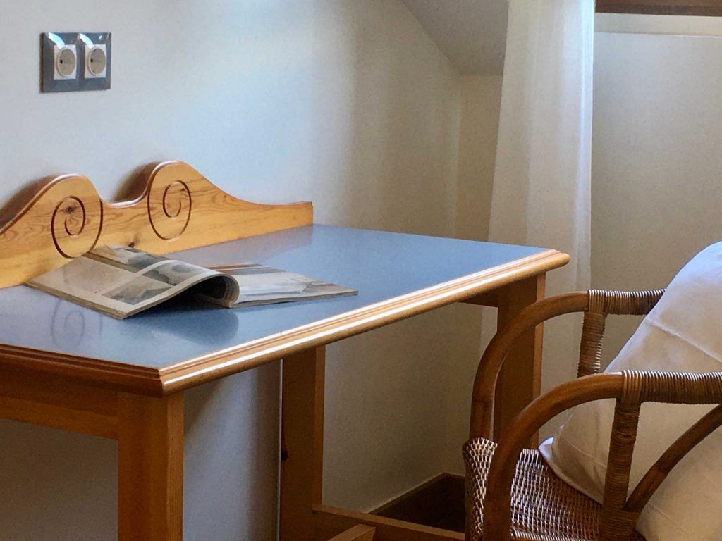 Desk with a blue surface, ornate wooden headboard, and wicker chair, near a window with curtains; a magazine lies on the desk.