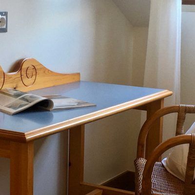 Desk with a blue top, wooden frame, and a magazine. A wicker chair is to the right.
