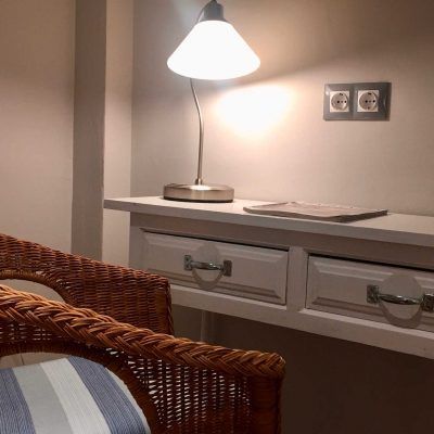 Wicker chair next to a white table with a lamp and electrical outlets on the wall.