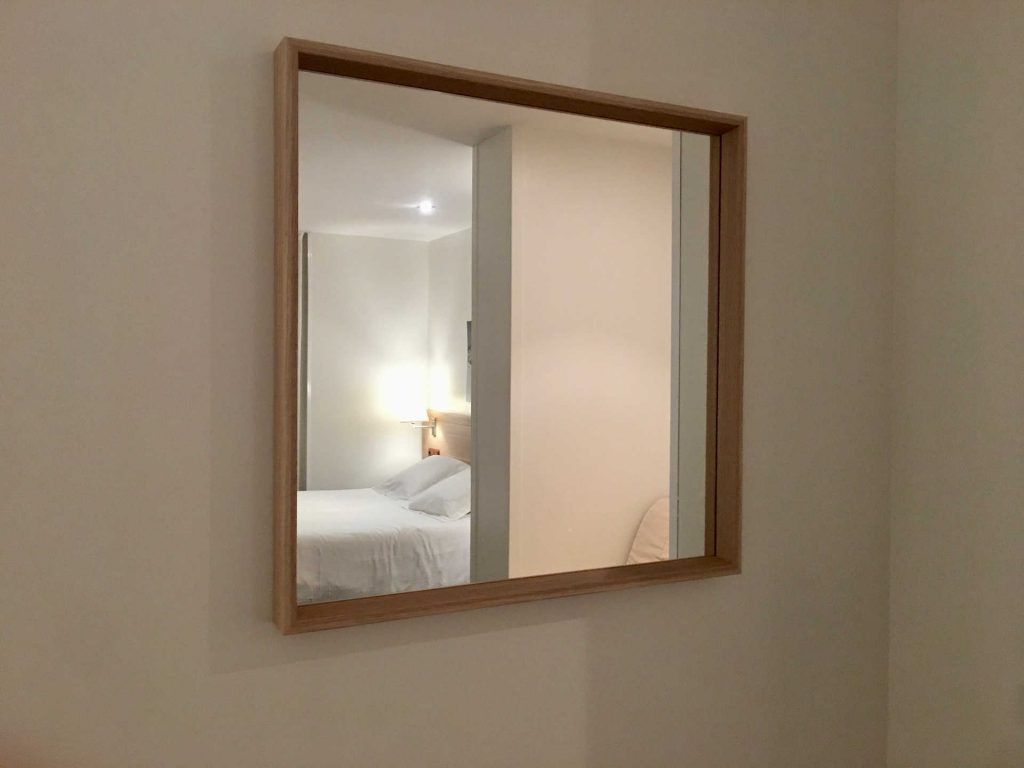 Square mirror with wooden frame reflecting a bedroom: white bedding, lamp, and walls.