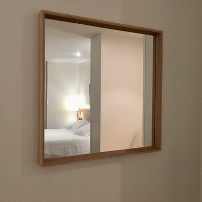 Square mirror with wooden frame reflecting a bedroom.