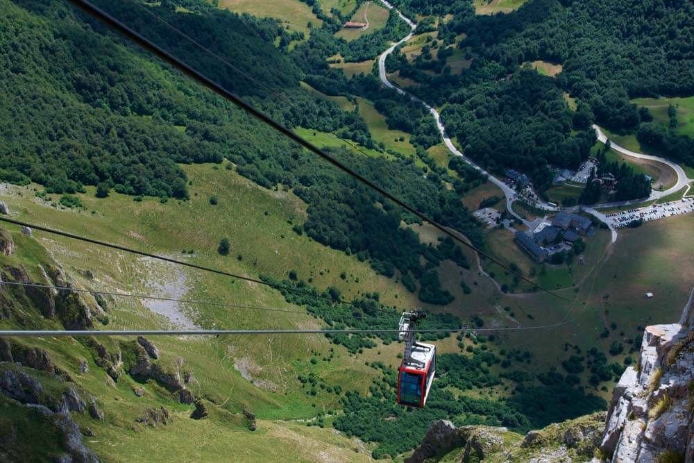 Cable car descending a steep, green valley with forests and a road visible.