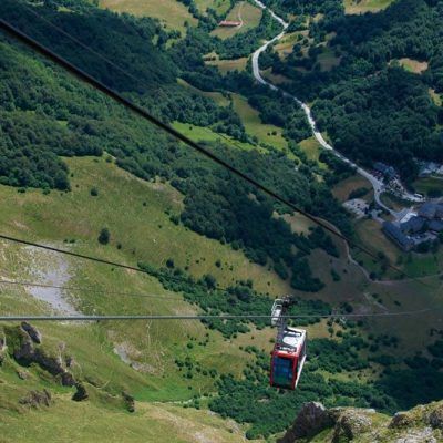 Cable car descending a green mountain valley. A road winds below near buildings.