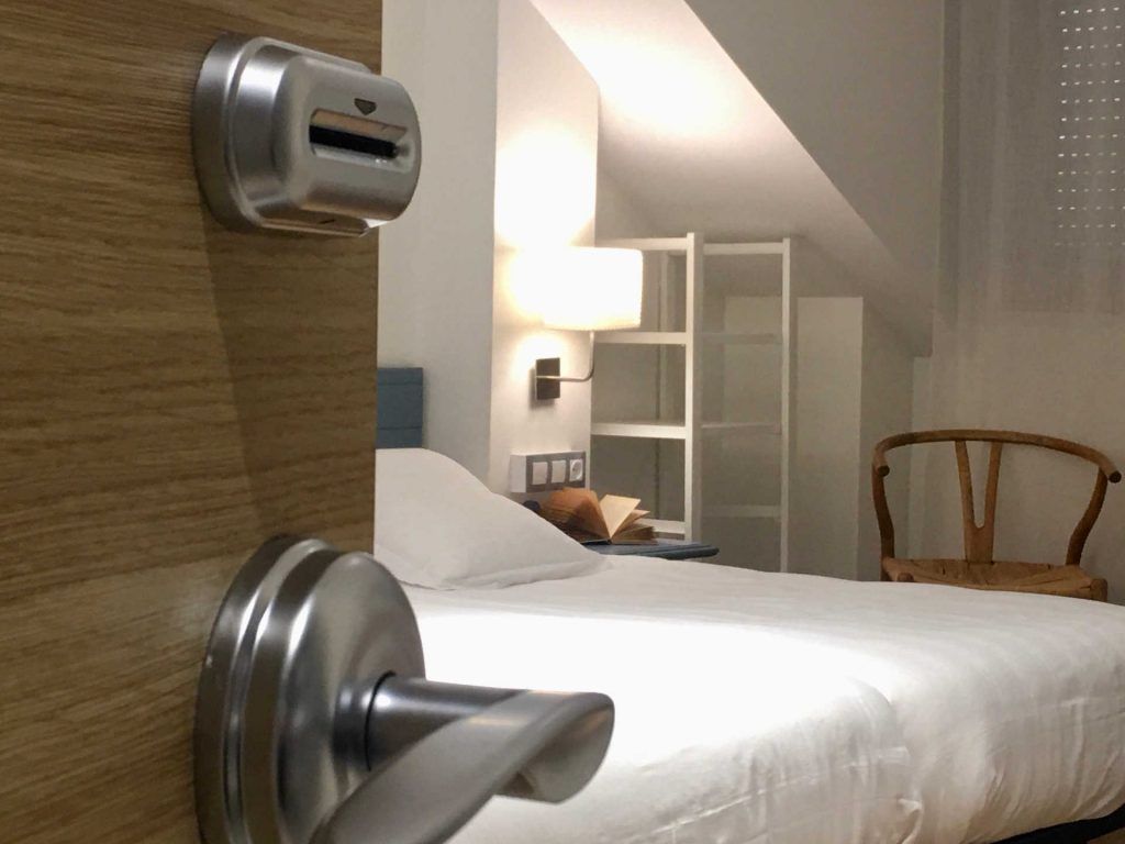 Door with card reader and handle, opening into a hotel room with bed, chair, and shelves.