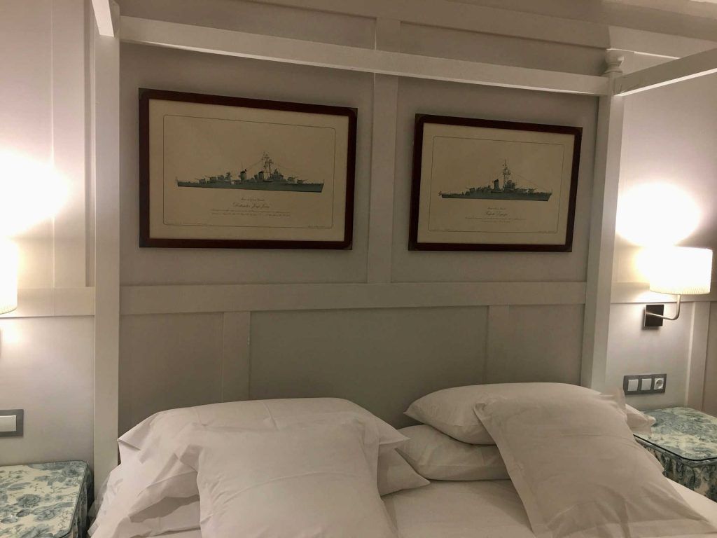 Bedroom with two framed ship diagrams above a bed with white linens and pillows.