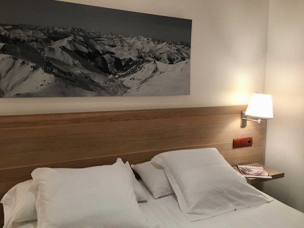 Bedroom with white bedding, wooden headboard, mountain print, and bedside lamp.