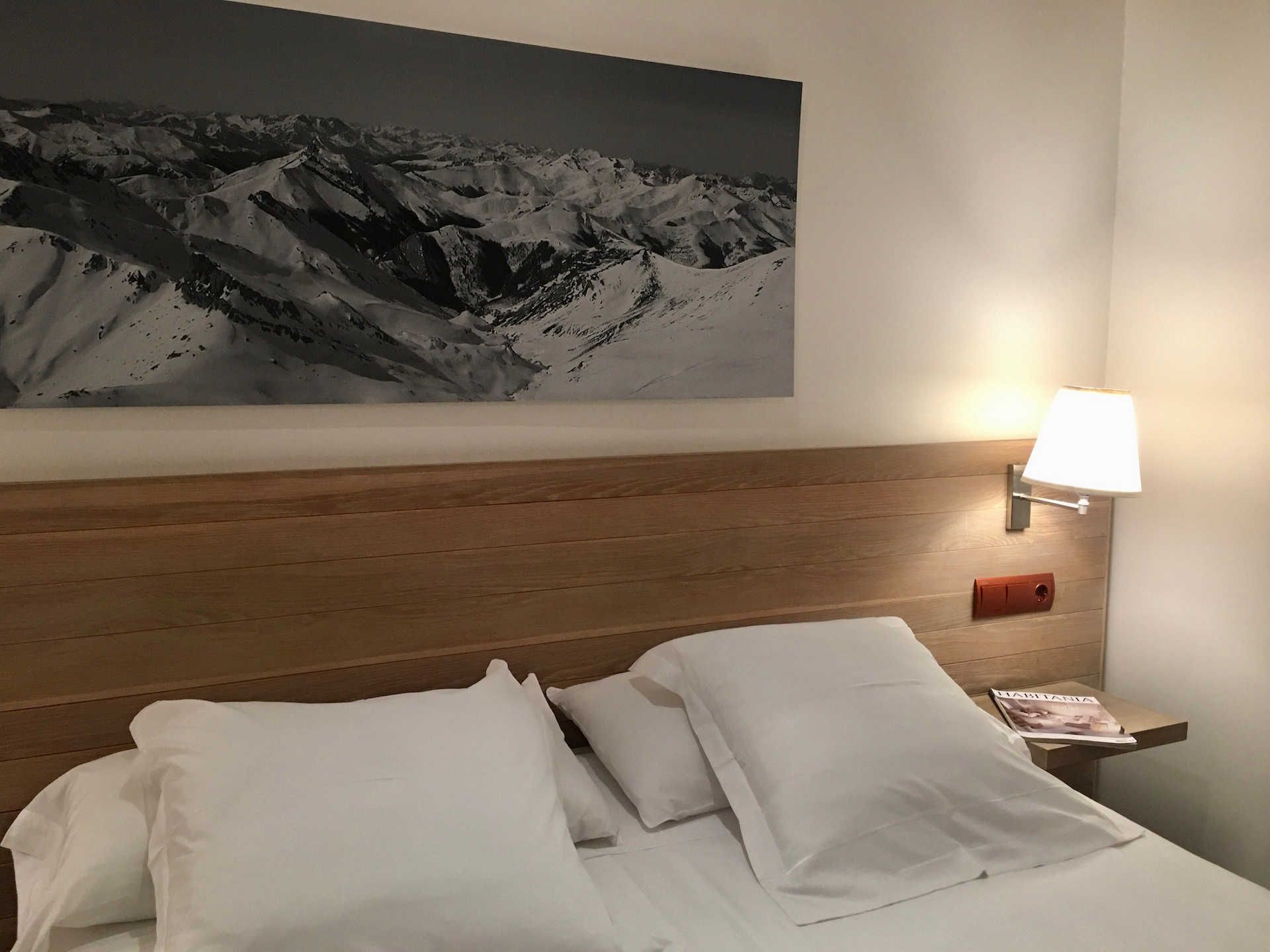 Bedroom with a double bed, white linens, wooden headboard, mountain artwork, and a bedside lamp.