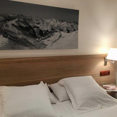 Bedroom with a large bed, pillows, and a snowy mountain landscape art piece. A lamp is lit.