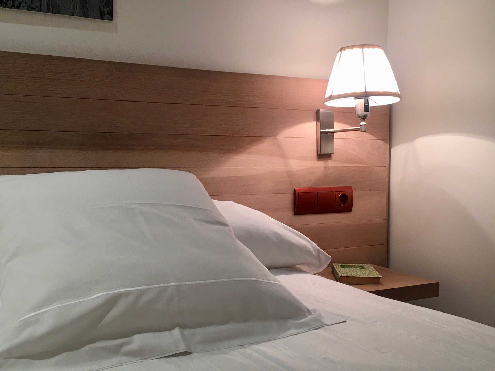 Bed with white linens, lamp on headboard, small nightstand, and light wood headboard.