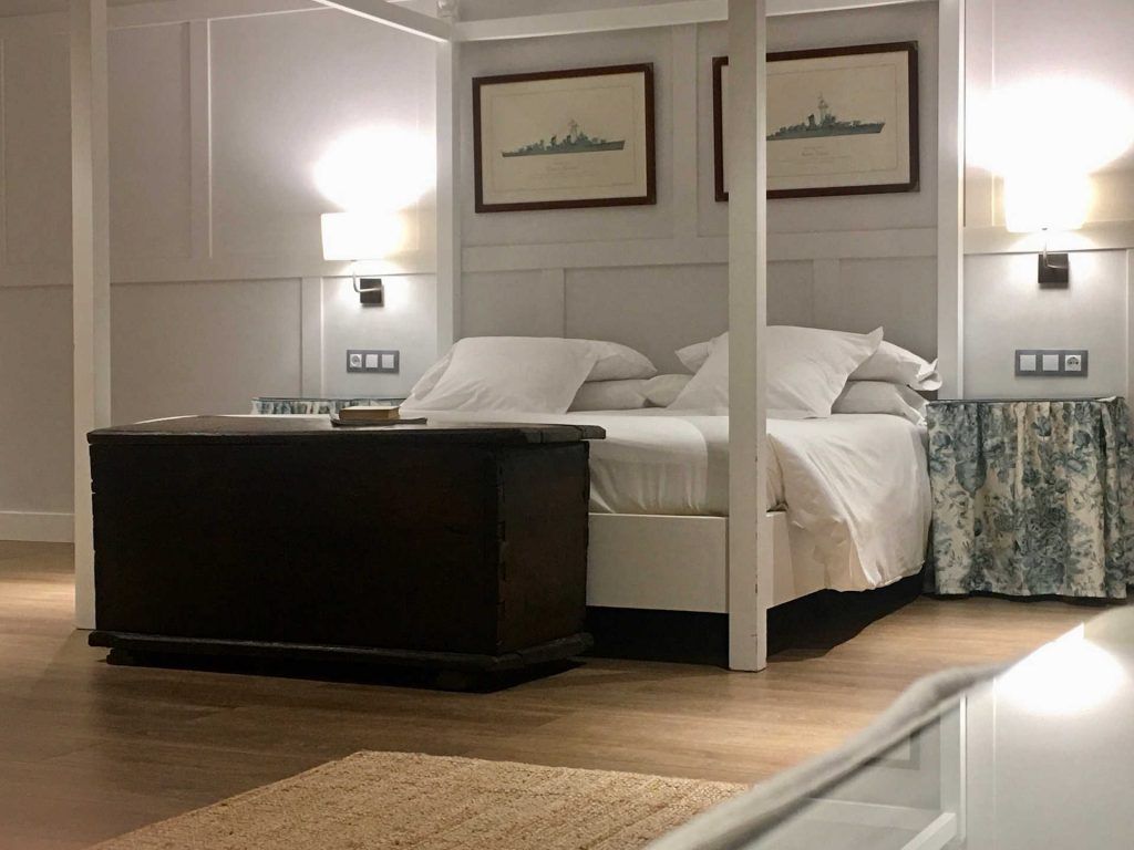 Bedroom with a white canopy bed, two framed ship blueprints, bedside lamps, and a wooden trunk.