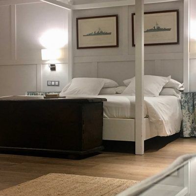 Bedroom with white canopy bed, dark wooden trunk, nautical art, and wood floors.