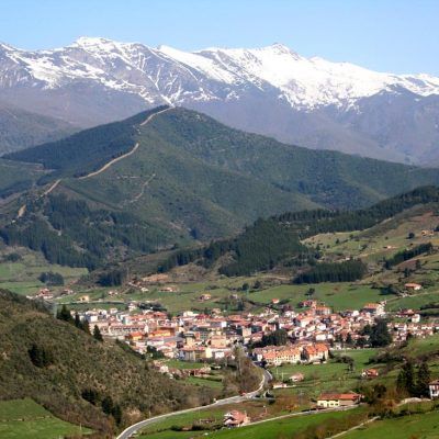 A village nestled in a valley with snow-capped mountains in the background. Green hills and blue sky.