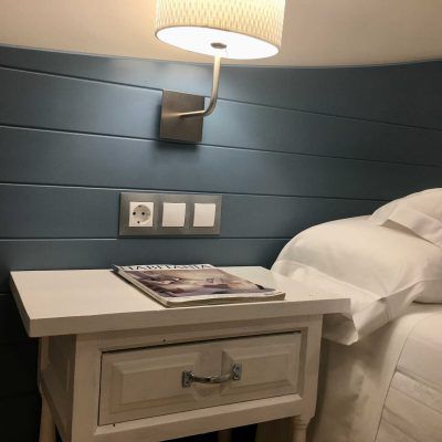 White nightstand with magazine, lamp, and bed next to blue paneled wall with electrical outlets.