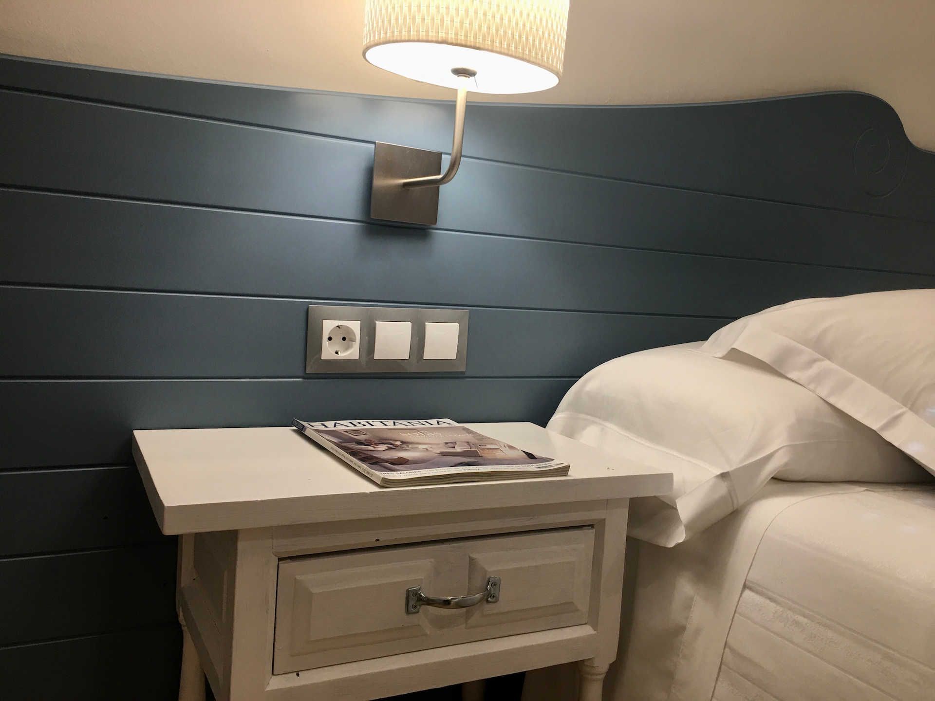 Nightstand with a magazine, beside a bed and wall lamp, against a blue paneled wall.