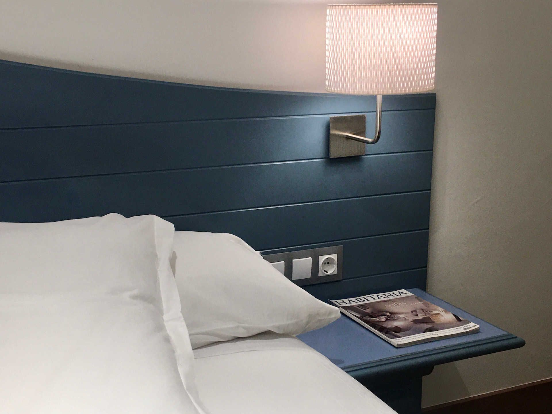 Bed with blue headboard, lamp, nightstand with magazine, and white linens.