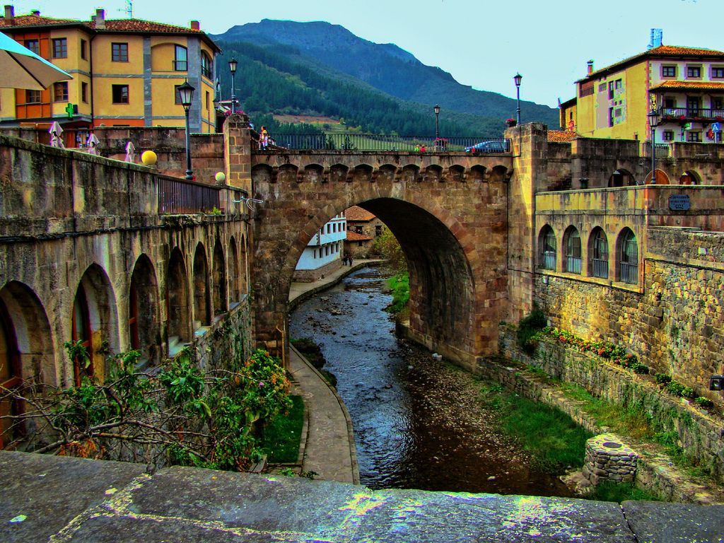 Stone bridge with arch over a river, connecting buildings in a European town, mountain backdrop.