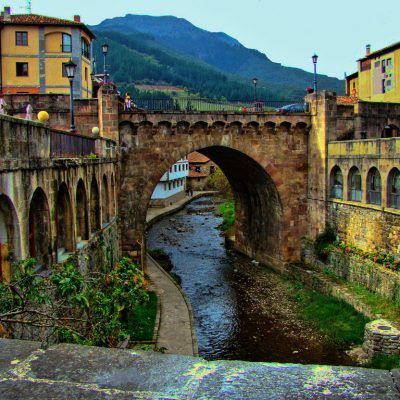 Stone bridge over a river in a mountain village, with buildings on both sides and a lush green mountain in the background.
