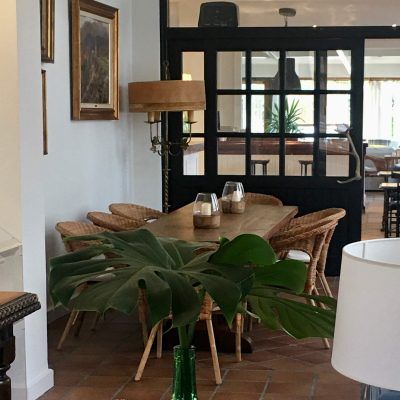 A dining area with a wooden table, woven chairs, and a large plant in front. A black-framed doorway leads to another dining space.