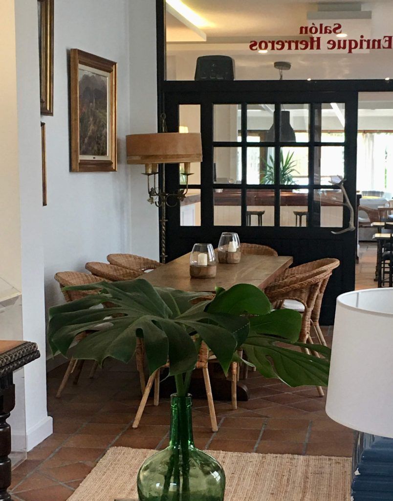 Dining area with wooden table, woven chairs, and a large green plant. A doorway leads to another room.