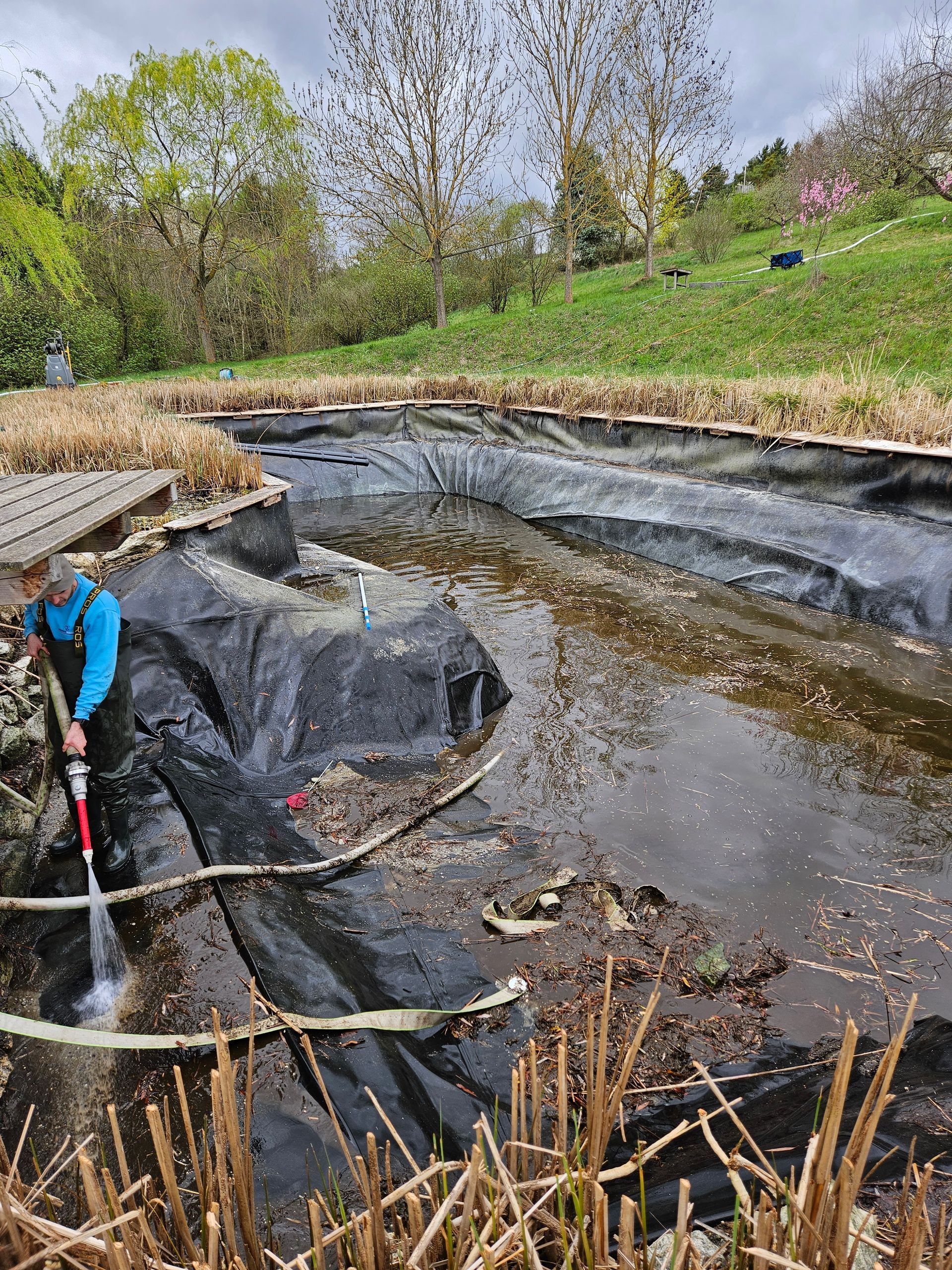 Eine Person spritzt Unrat aus einem Teich. Der Teich hat eine schwarze Teichfolie.