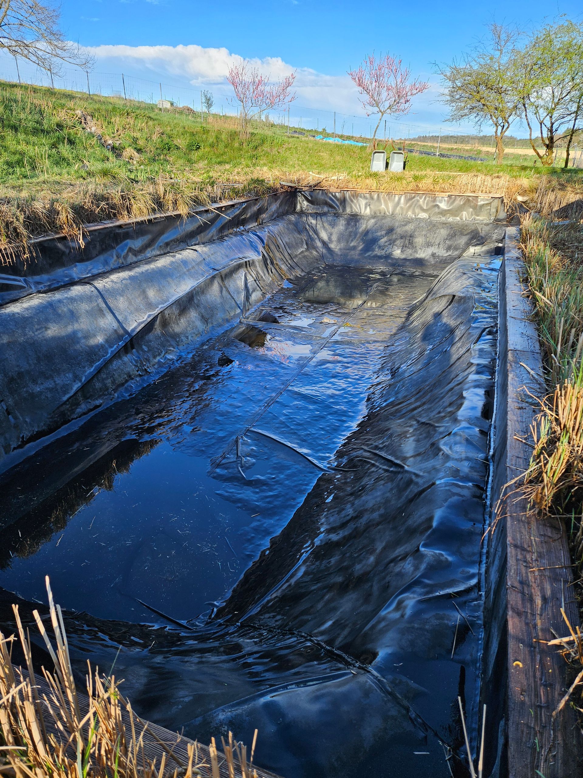 Schwarz umrandeter Wasserspeicher mit gewellter Oberfläche, eingebettet in eine grasbewachsene Landschaft.