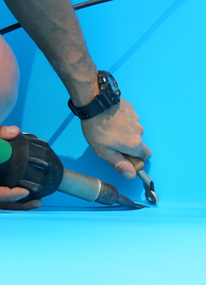 Person, die mit einer Heißluftpistole und einem Handwerkzeug blaues Material bearbeitet.