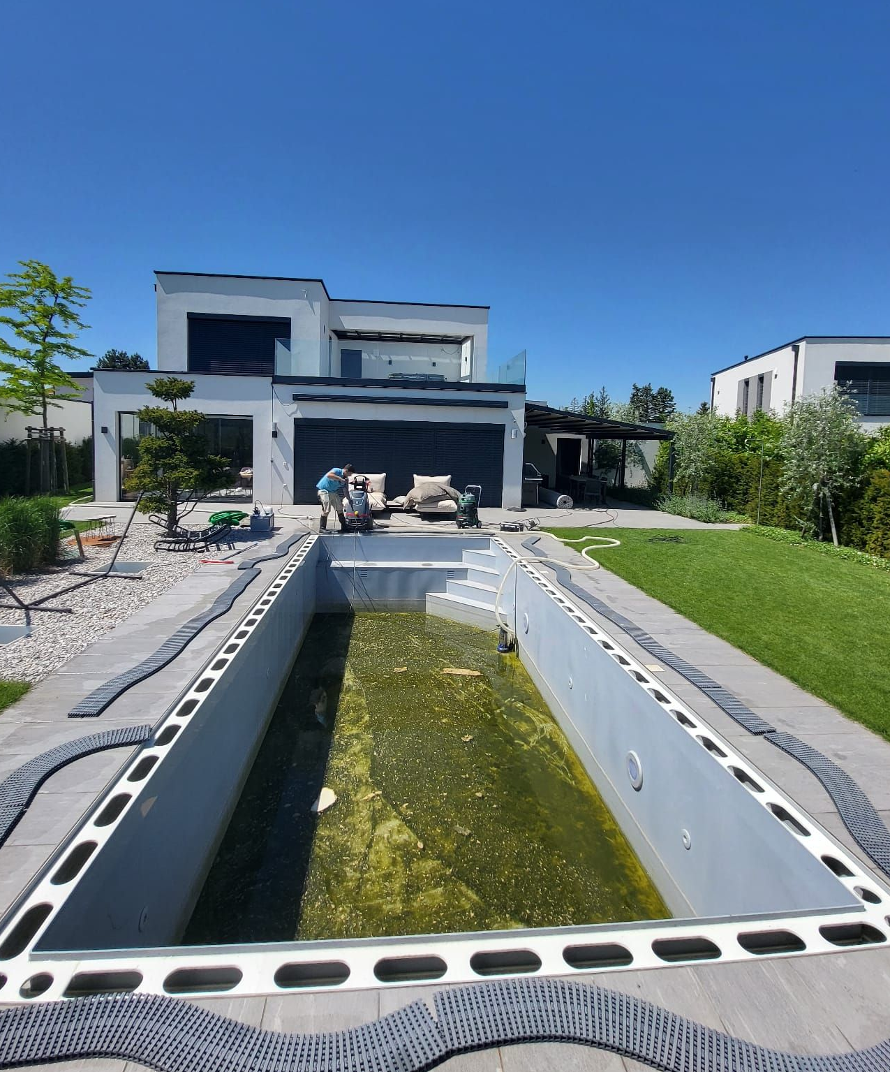 Ein leerer, mit Algen bedeckter Swimmingpool, arbeitende Männer, im Hintergrund ein modernes Haus an einem sonnigen Tag.